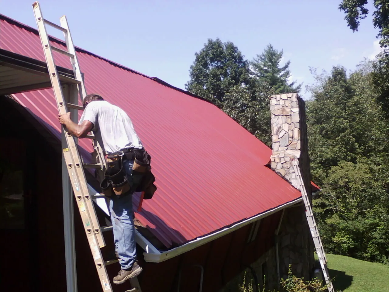 Roofer on a ladder beside a red metal roof and stone chimney — Soffit & Fascia Repair by Roofing Brothers in Cookeville